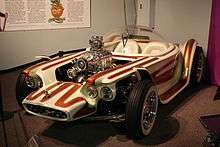 A white car with brown stripes, with open wheels and a clear bubble canopy over twin seats, and exposed, chromed engine with a blower. Beatnik Bandit II and a few of Roth's other cars are also on display in this museum.