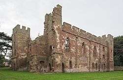 A ruined building minus a roof and with crumbling walls. Some of the walls are crenelated. The walls are build with red stones in the middle and grey stones as edging on the tops and corners.