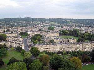 Distance photograph showing rows and crescents of yellow stone buildings. Several tress and hills in the background.