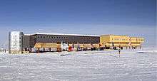 A long, large building consisting of several sections stands behind a line of flags flying on poles. The ground surface is ice-covered; in the middle foreground is a short striped pole which indicates the position of the South Pole