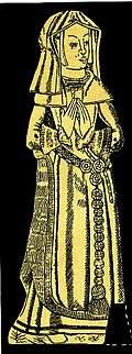 A woman in a long gown standing with hands clasped prayer-like in front of her. She wears a gable headdress and veil, a fur-lined gown over a kirtle with a girdle or belt. There are large paternoster beads attached to her decorative belt by a three-rose buckle.