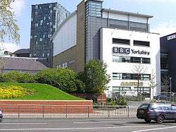 A quirky modern five-storey building with a large sign saying "BBC Yorkshire" in black above the second-floor windows on the white-fronted façade of the lower four floors can be seen on the far side of a dual-carriageway road with a barrier along the central reservation. At right-angles to the right of the building is a tall blue slab with the letters "BBC" in white at the top. The left side of the building is mostly brick-red with a few windows, but above it is a light blue windowless section. The roof above this and the grey fifth floor of the frontage curves gently down to the rear. A lone car is driving from left to right along the road; between it and the building, temporary boards have been erected in front of a building to the left. In the top left-hand corner of the picture, part of a tall many-windowed building can be seen.