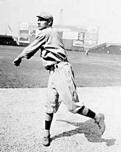 A man in a white baseball uniform and white baseball cap follows through after throwing a baseball on a grass field.