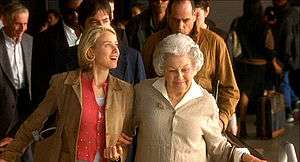 Naomi Watts beaming and facing into soft light holding the arm of an older woman while they take a down escalator at Los Angeles International Airport