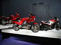 Three sporty motorcycles of the 1990s on pedestals. Text on the wall behind them says "1982-1989" with a few dozen words relevant to the period, such as "Atari", "MTV", and "Robert Mapplethorpe".