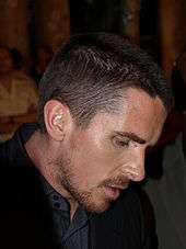 Profile shot of a man looking down and to his right. He has a goatee and stubble, and is wearing a black dress shirt with collar and a black blazer.