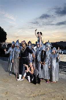 Chrome Hoof on a dawn/dusk Summer of 2007 river beach, dressed to promote 'Pre-Emptive False Rapture'. Flanking members hold candelabra and most are wearing silvery cowls, with Shingai Shoniwa counterpointed in matt black.