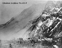 A small town shown at the base of a mountain. The mountain's face stands barren following a large rockslide and a light cloud of dust is visible in the air.