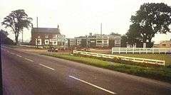 A faded period photograph of the motel on a grey day. The main road is in the foeground, from lower right to half way up the left side. In the middle distance a two-story 18th or 19th century building stands at right angles to the road, it's windowed frontage facing our vantage point. It is on the opposite side of the road to us, beyond the entrance track. This side of the track, in the centre of the photograph and still on the opposite side of the road to us are a range of low, flat-roofed, single story buildings, almost certainly new at the time. There is low whir post-and-rail fencing on the verge this side of the motel. The older building is probably red brick. The new buildings have dark grey walls. The sign reading 'Trenton Motel' is clearly visible by one of the modern buildings.