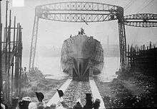 A black and white photograph showing a ship's hull with no superstructure sliding down a slipway into a waterway