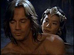 Upper body shot of a bare-chested muscled man with shoulder length brown hair. Behind him is an Asian woman with long black hair, wearing an ornate headdress.