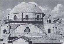 A large domed roof sits upon a bricked parapet, which includes arched windows, three of which are visible. The base of the parapet is surrounded by a narrow veranda, which is fenced with decorative metal grating. Below this, a central clover shaped window lies at the top of an arched wall. Two supporting towers stand on the left and right, each containing an arched window. The right tower, capped with a small dome, is a storey higher, reaching to the top of the bricked parapet, which supports the domed roof.