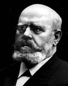 Head and shoulders of a middle-aged man looking with dignity to the left of the viewer. He is bald, has a short, but stout, beard, and is wearing small glasses and a tie-less suit.