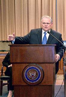 Clean-shaven man in his 60s, with gray hair, wearing glasses, dressed in a dark suit and blue tie, speaking from behind a dark, varnished wooden lectern, with his right arm outstretched. The front of the lectern is emblazoned with the Great Seal of the United States.