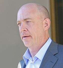 A middle-aged man with close-cropped hair and wearing a gray jacket with a sky blue shirt faces left