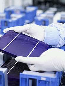 A part of gloved hands are hold a tablet-sized solar cell with three panels.
