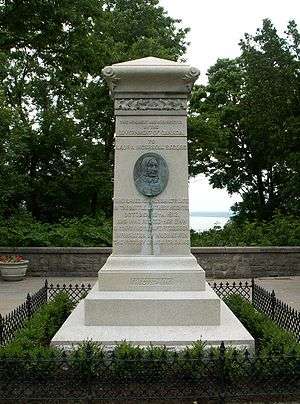 An upright stone monument surrounded by a low hedge. In the middle is an upright oval of an elderly woamn's face in relief.