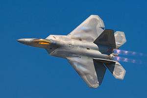 A pilot peers up from his F-22 Raptor while in-flight, showing the top view of the aircraft. The terrain of Nevada can be seen below mostly cloudless skies. Aircraft is mostly gray, apart from the dark cockpit canopy.