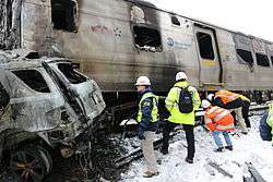 Several people in neon colored safety clothing and white helmets examine the fire-damaged wreckage of a silvery passenger rail car along the top with "Metro-North Railroad" written on it. One person has the letters "NTSB" on their jacket sleeve. At left is the rear of a similarly fire damaged automobile.