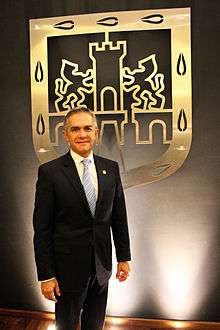 Miguel Ángel Mancera stands in front of a golden shield depicting an old Mexico City's shield. He looks directly to the camera. He wears a black suit.