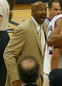 A man, wearing a brown suit and white shirt, is shouting while standing on a basketball court, surrounded by several other people.