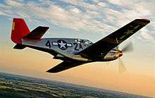 A single nosed-propeller aircraft is in mid-flight above land. The red nose faces right and the underside of the aircraft is slightly in view. The aircraft has black propellers, gray base paint, and black letters reading: "A42". The "4" and "2" are separated by a United States roundel in black with a central white star. The roundel is also visible on the tops of the wings. The full length of the right side of the aircraft is visible.