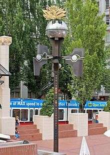 A color photograph of an urban center area. A tall, thin structure stands adorned with various lights and appendages, with a globe-like object at the top.