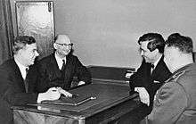 Four men sit facing each other around a table. Three of the men wear civilian suits, while one wears a military uniform