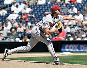 A left-handed, middle-aged white male baseball pitcher, wearing a grey uniform and a red cap, in a throwing stance.