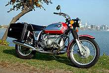 Red BMW R75/5 motorcycle fitted with panniers, parked on grass by a lake with a city in the background