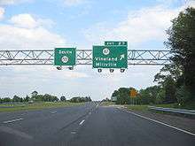 A four lane freeway at an interchange. Two green overhead signs stand over the road with the left one reading south Route 55 and the right one reading exit 27 Route 47 Vineland Millville with an arrow pointing to the upper right
