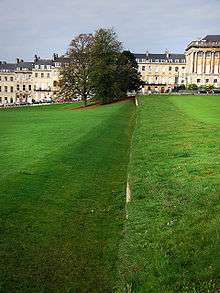 A ditch or trench in the grass with the right hand wall vertical and the left hand wall sloping. In the background are trees and yellow coloured stone buildings.