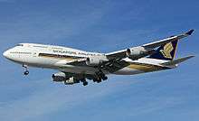 A large mostly-white four-engine jet airliner with golden stylized bird design, on approach towards left of screen with landing gear extended