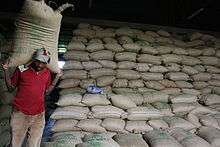 A man carries a large bag on his shoulders. Behind him, thousands of bags are stacked in rows.