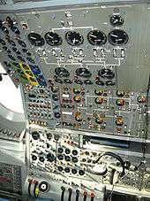 An airliner flight deck control panel with many instruments and switches. A pair of headphones hang from the panel.