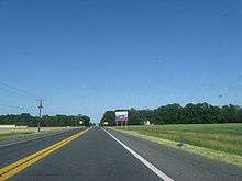 A two-lane road passing through farm fields.  A blue and white sign on the right indicates an ongoing construction project on US&nbsp;113
