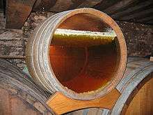 Photo showing a barrel with a glass bottom revealing the layer of yeast on the surface under which the yellow wine is ageing. This yeast is in the form of an irregular white layer - it forms small stalactites that sink a few millimetres into the wine which is already dark yellow wine. The wall in the background is grey, probably coated in grey mould.