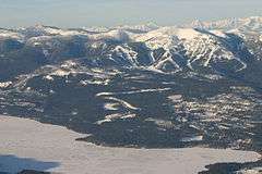 Aerial view of a forested mountainous area, with ski trails. In the foreground is a partially frozen body of water.